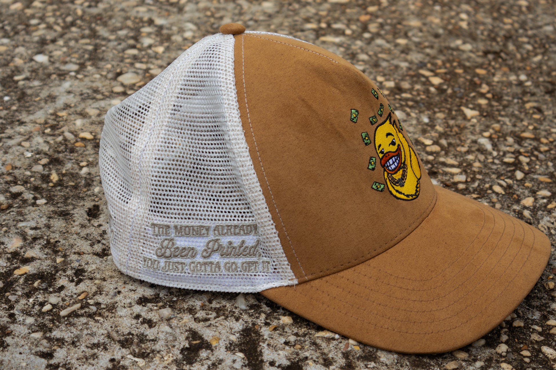 Brown and white cap with a yellow duck design on a concrete surface. Embroidery on the side of the hat says "The money already been printed, you just gotta go get it
