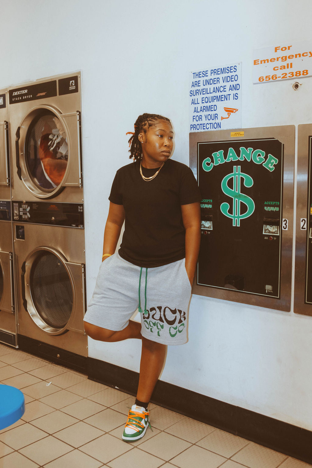 Person standing in a laundromat with washing machines and a sign on the wall wearing grey shorts and a black and green design on the leg.